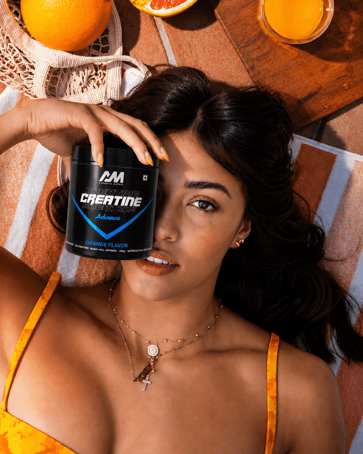 A woman lying on a striped beach towel holds a black tub of CMA Creatine Advanced powder near her face. Orange slices and a glass of orange juice sit on a wooden table in the background.
